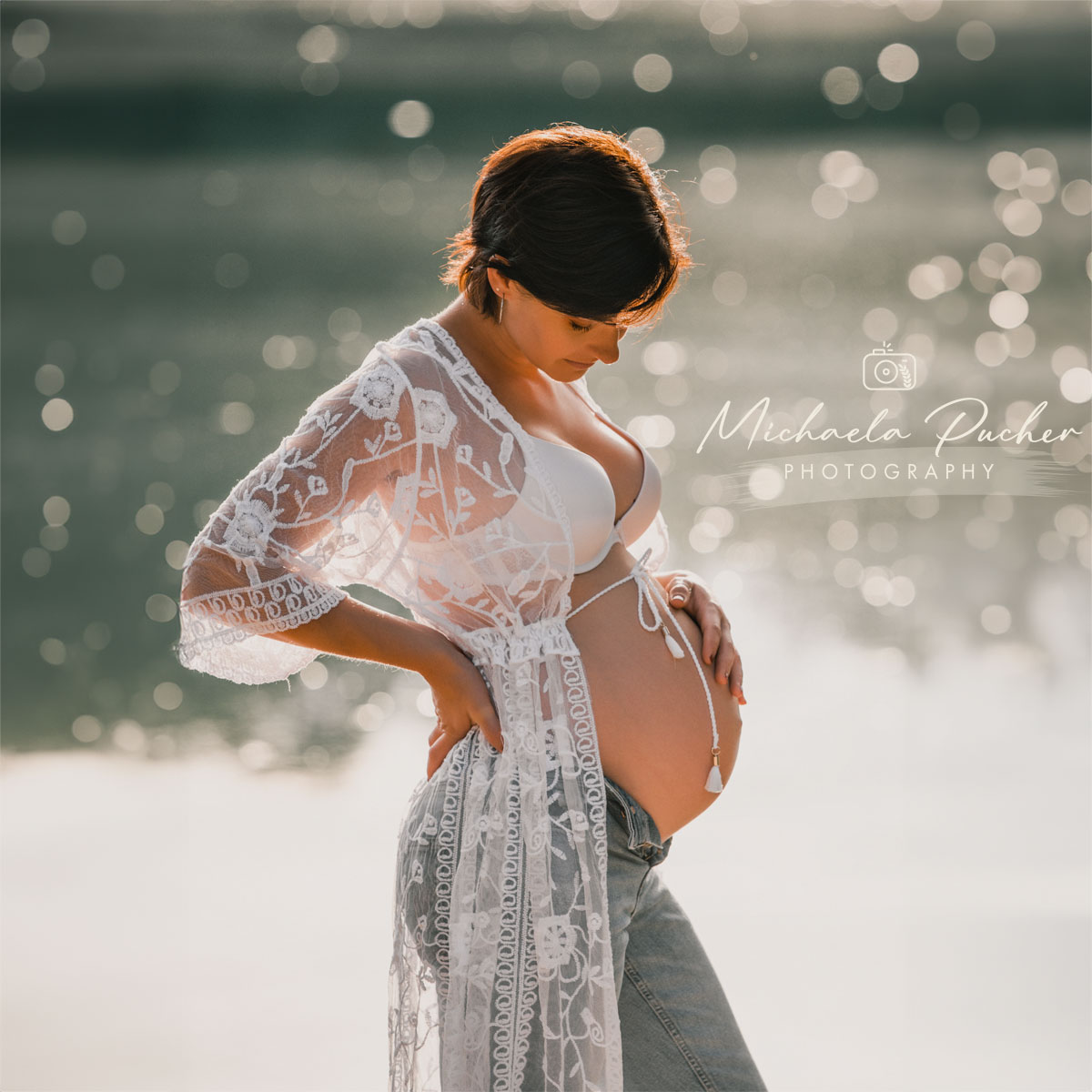 Schwangere Frau steht am Wasser im Gegenlicht, trägt eine bestickte weiße Spitzenrobe über Jeans und BH und hält liebevoll ihren Babybauch, während das Licht im Hintergrund glitzert.
